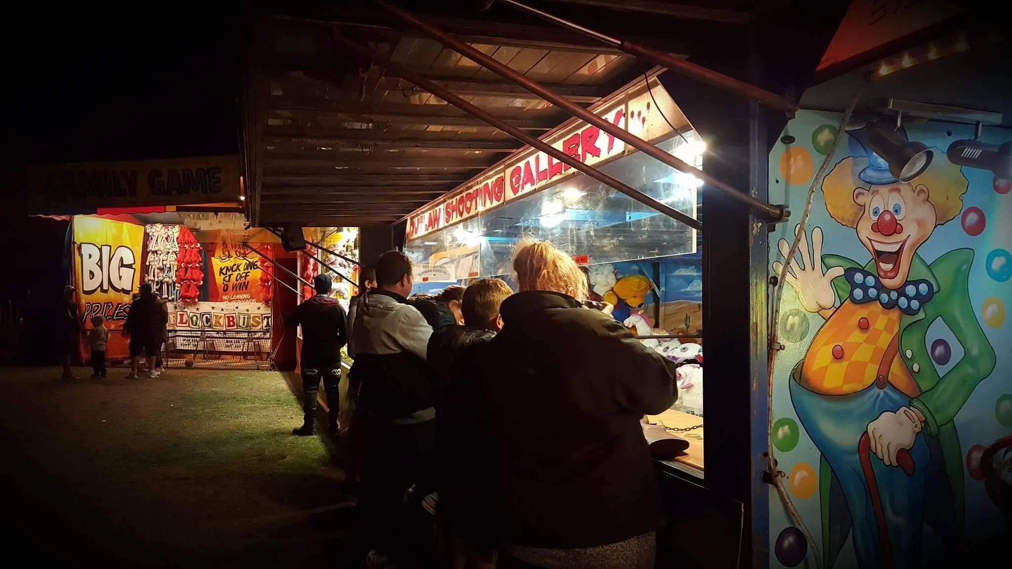 Visitors at a game stall at night beside a painted clown mural